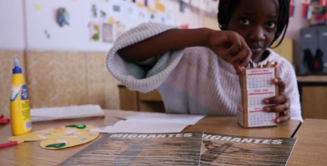 Una niña trabaja en un taller de manualidades junto a ejemplares de la revista MIGRANTES sobre la mesa