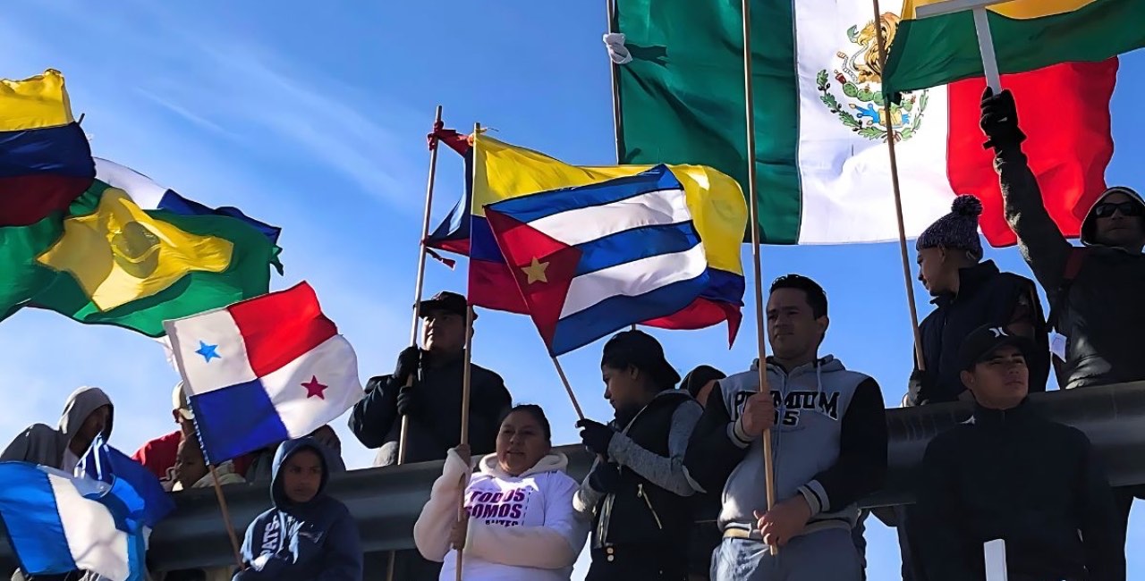 Personas migrantes ondean banderas de México, Cuba, Colombia, Panamá y otros países bajo un cielo despejado