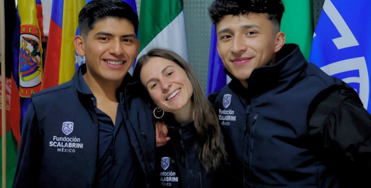 Tres voluntarios con chamarras de la Fundación Scalabrini sonríen frente a las banderas de distintos países